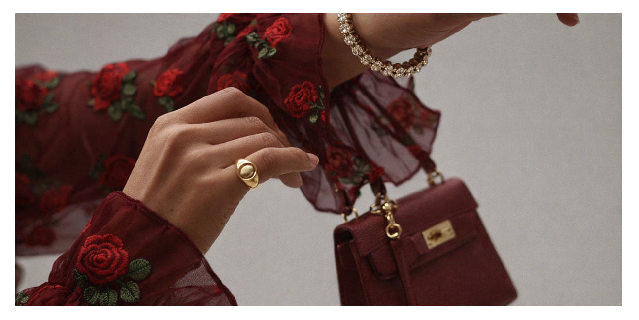 Hand holding a burgundy handbag with gold accents, wearing a matching ring and bracelet.